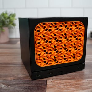 Könnte beinhalten: Ein schwarzes, rechteckiges elektronisches Gerät mit einer markanten orangefarbenen Frontplatte, die ein komplexes, sich wiederholendes wellenförmiges Gittermuster aufweist. Die Basis des Geräts enthält zwei sichtbare USB-Anschlüsse.