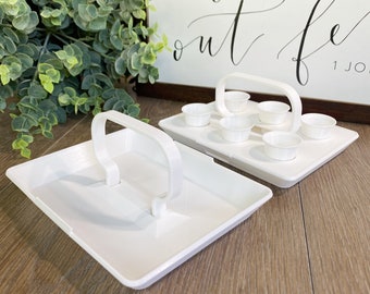 May include: White plastic tray with a handle, divided into six sections, each with a small white cup. The tray is open and the cups are empty. The tray is on a wooden surface.