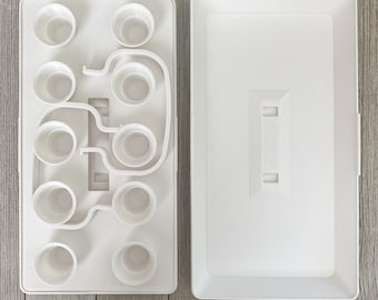 May include: A white plastic paint palette with multiple circular wells and a separate lid. The palette has a unique design with interconnected wells of varying sizes, ideal for holding different colors of paint or other art supplies.