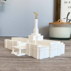 May include: White miniature replica of a temple with a gold angel moroni statue on top. The temple has a white steeple and a white entrance with columns.