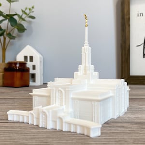 May include: White 3D printed model of a temple with a gold statue on top of the spire. The model is sitting on a wooden surface.
