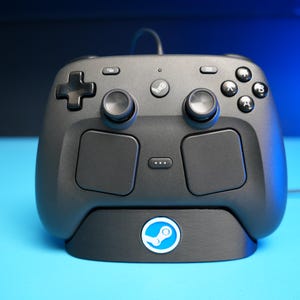Può includere: Un controller per videogiochi grigio scuro dotato di due levette analogiche, un pad direzionale, quattro pulsanti azione etichettati A, B, X e Y, due ampi touchpad e un pulsante centrale con il logo Steam, posizionato su una base coordinata.