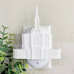 May include: A white architectural night light shaped like a temple. The night light has a detailed facade with columns, windows, and a tall spire. The night light is plugged into a white outlet.