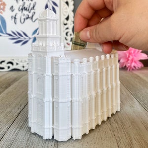 May include: A white architectural model of a temple with a domed top and multiple towers. A hand is inserting a bill into a slot on the roof. The model is on a wooden surface.