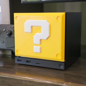May include: A black computer case featuring a yellow front panel with a raised white question mark symbol. The front panel includes circular indentations at each corner. A black game controller is positioned to the left of the square case.