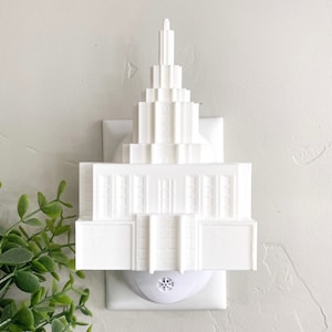 May include: White ceramic nightlight shaped like a building with a spire. The nightlight plugs into a standard electrical outlet.