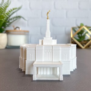 Puede incluir: Modelo 3D impreso en blanco de un templo con una estatua dorada en la parte superior. El templo tiene una gran entrada con columnas y un pequeño porche.