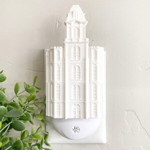 May include: A white architectural night light shaped like a building with multiple stories and a tower. The night light has a rectangular base and a decorative design. The night light is plugged into a white wall.