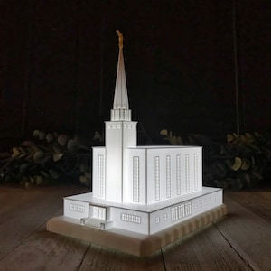 May include: A white 3D printed model of a temple with a gold spire. The model is lit from within, creating a warm glow.