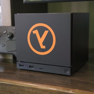 Op de afbeelding: Een matzwarte, kubusvormige gameconsole met een opvallend oranje cirkelvormig logo met een gestileerde letter Y in het midden. Het voorpaneel bevat twee USB-poorten, een klein indicatielampje en een ronde aan-uitknop rechtsonder.