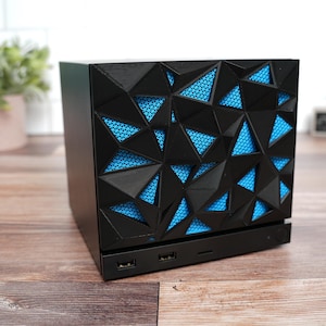 Puede incluir: Una caja de ordenador negra con un panel frontal geométrico y facetado con una malla azul brillante detrás. La base de la unidad incluye dos puertos USB y un botón de encendido. El diseño consta de formas triangulares angulares que crean una apariencia texturizada y en capas en la superficie frontal de la carcasa rectangular.