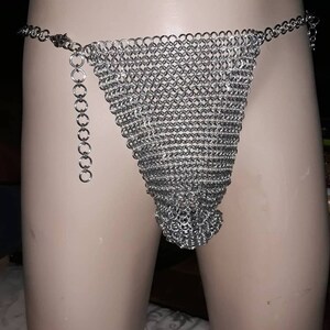 Peut inclure: Un string en cotte de mailles argentée, avec un devant triangulaire et une ceinture en chaîne. Le string est fait de petits anneaux métalliques imbriqués, créant un vêtement souple mais structuré. Une chaîne décorative pend sur le côté.