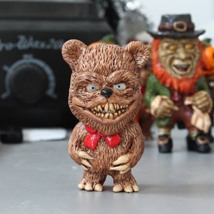 Puede incluir: Una figurita de oso de peluche marrón con una pajarita roja y una sonrisa amenazante. El oso tiene garras afiladas y una textura peluda.
