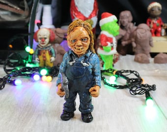 May include: A hand-painted figurine of a horror movie character wearing a blue and white plaid shirt, blue overalls, and holding a small axe. The figurine has a creepy, realistic expression and is standing on a wooden surface.