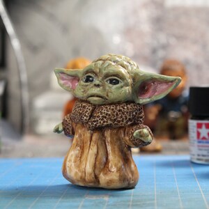 Puede incluir: Una peque&ntilde;a figura verde y marr&oacute;n de un personaje de la franquicia Star Wars. La figura lleva una t&uacute;nica marr&oacute;n y tiene orejas grandes.