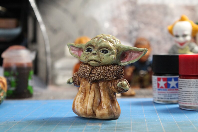 Puede incluir: Una peque&ntilde;a figura verde y marr&oacute;n de un personaje de la franquicia Star Wars. La figura lleva una t&uacute;nica marr&oacute;n y tiene orejas grandes.