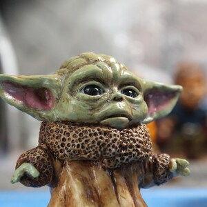 Puede incluir: Una peque&ntilde;a figura verde y marr&oacute;n de un personaje de la franquicia Star Wars. La figura tiene orejas grandes y un cuello de piel marr&oacute;n.