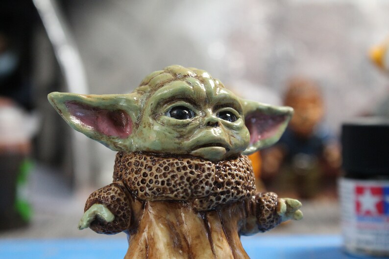Puede incluir: Una peque&ntilde;a figura verde y marr&oacute;n de un personaje de la franquicia Star Wars. La figura tiene orejas grandes y un cuello de piel marr&oacute;n.