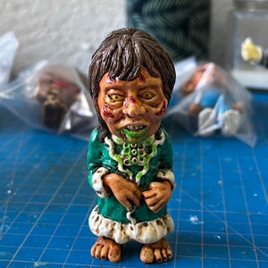 May include: A small green and white figurine of a person with a green face and yellow eyes. The figurine is wearing a green robe with white trim and has a green snake around its neck.
