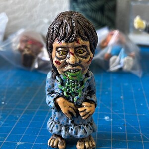 May include: A hand-painted ceramic figurine of a girl in a blue and white polka dot nightgown. She has green and brown eyes, a green and white mouth, and a green and white creature on her neck.