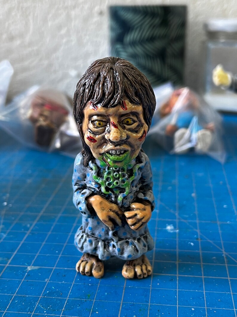 May include: A hand-painted ceramic figurine of a girl in a blue and white polka dot nightgown. She has green and brown eyes, a green and white mouth, and a green and white creature on her neck.