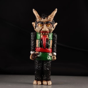 Op de afbeelding: Een Krampus-beeldje met een gedetailleerd, handgeschilderd ontwerp. De figuur heeft een dreigende uitdrukking met hoorns, een lange rode tong en een groen en rode outfit. De figuur staat rechtop.
