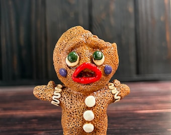 May include: A brown gingerbread man figurine with a distressed look. The figurine has green eyes, a red mouth, and white buttons. The figurine is standing on a wooden surface.