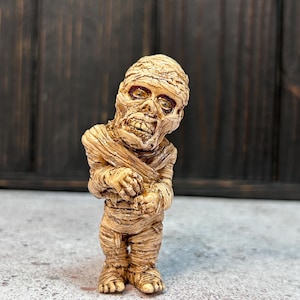 Puede incluir: Una pequeña figura de momia beige con una cara detallada y un cuerpo envuelto. La figura está de pie sobre una superficie gris claro.