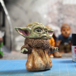 Puede incluir: Una peque&ntilde;a figura verde y marr&oacute;n de un personaje de la franquicia Star Wars. El personaje tiene orejas grandes, un abrigo de piel marr&oacute;n y una expresi&oacute;n seria.