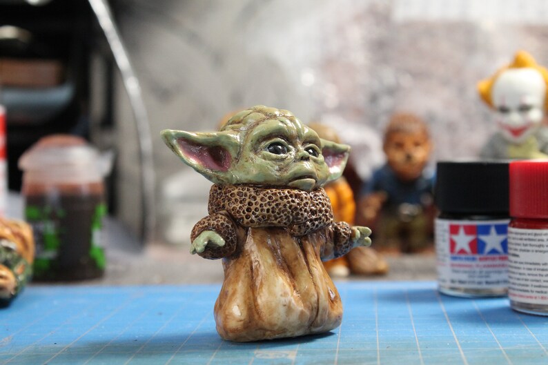 Puede incluir: Una peque&ntilde;a figura verde y marr&oacute;n de un personaje de la franquicia Star Wars. El personaje tiene orejas grandes, un abrigo de piel marr&oacute;n y una expresi&oacute;n seria.