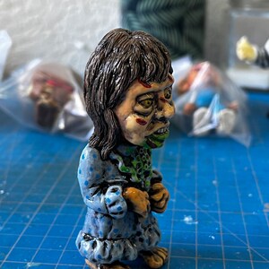 May include: A hand-painted figurine of a girl with brown hair, wearing a blue and white polka dot dress. The figurine has green and red paint details on the face and a green and white painted neck.