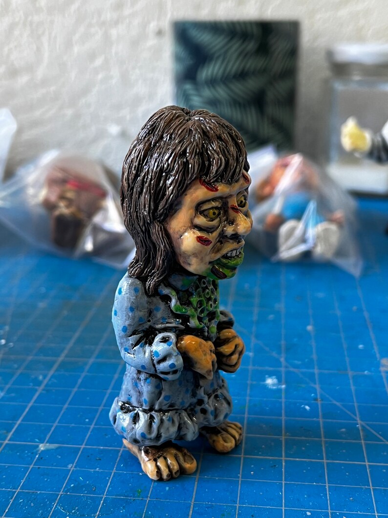 May include: A hand-painted figurine of a girl with brown hair, wearing a blue and white polka dot dress. The figurine has green and red paint details on the face and a green and white painted neck.