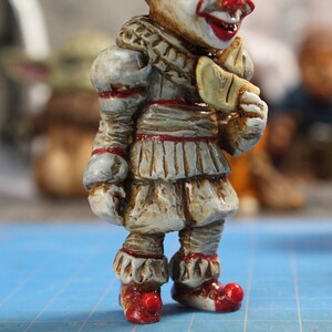 Puede incluir: Primer plano de una figurita pintada de Pennywise el payaso bailar&iacute;n de la pel&iacute;cula "IT". La figurita lleva un traje blanco y rojo con un cuello de volantes y zapatos rojos. La figurita sostiene un globo amarillo en su mano derecha.