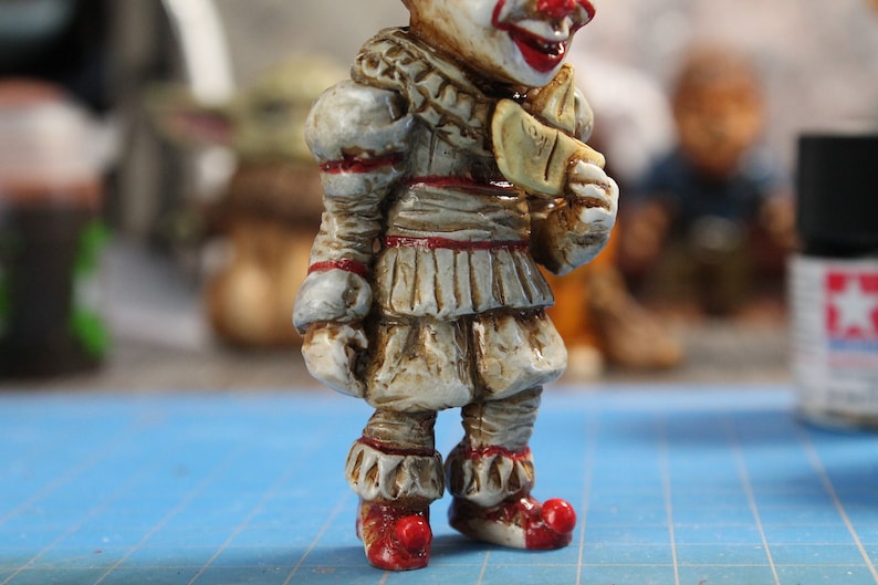 Puede incluir: Primer plano de una figurita pintada de Pennywise el payaso bailar&iacute;n de la pel&iacute;cula "IT". La figurita lleva un traje blanco y rojo con un cuello de volantes y zapatos rojos. La figurita sostiene un globo amarillo en su mano derecha.
