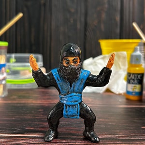Könnte beinhalten: Eine handbemalte Figur eines Ninja-Kriegers mit einem blauen Band und schwarzer Kleidung. Die Figur ist in Kampfstellung mit erhobenen Armen.
