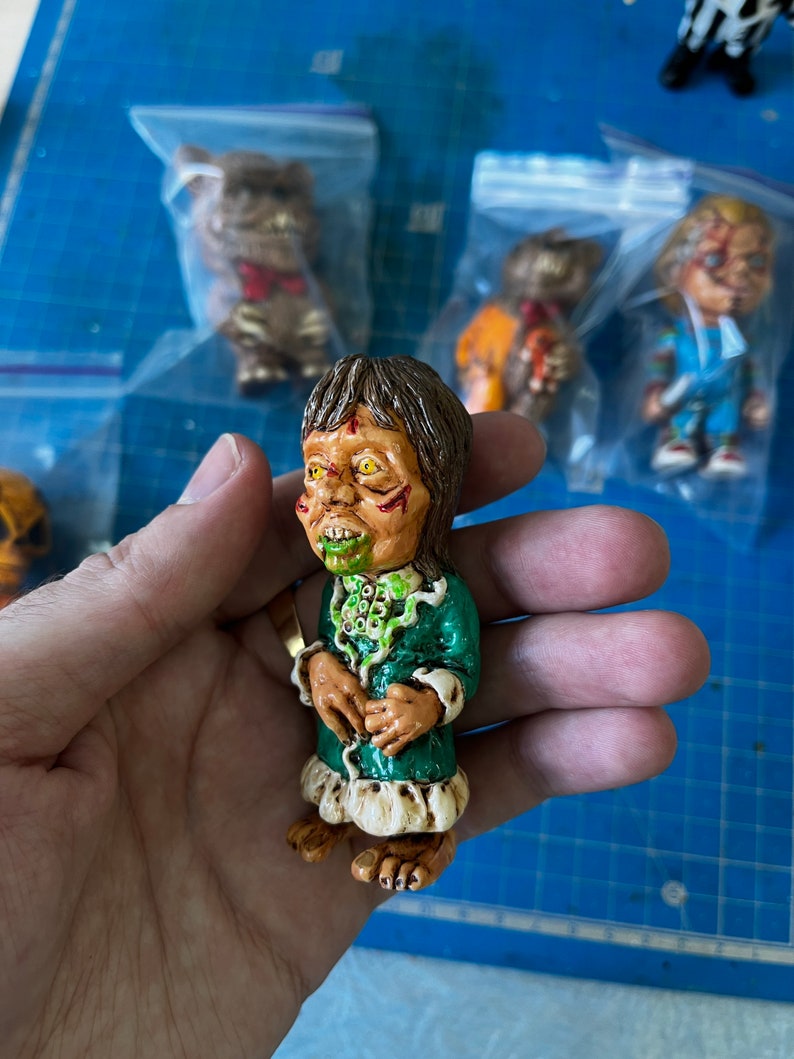 May include: A small green and white figurine of a monster with a green face and yellow eyes. The monster is wearing a green dress with white trim and has a green and white necklace.