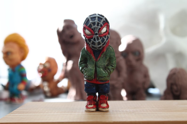 Puede incluir: Una peque&ntilde;a figura verde y roja de un superh&eacute;roe que lleva una m&aacute;scara negra y gris con un dise&ntilde;o de telara&ntilde;a. La figura est&aacute; de pie sobre una superficie de madera.