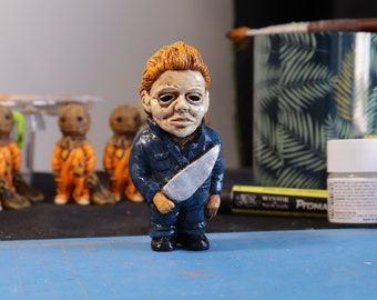May include: A hand-painted ceramic figurine of a horror movie character holding a silver knife. The figurine is wearing a blue jumpsuit and has a white mask with red eyes. The figurine is standing on a blue cutting mat.