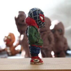 Puede incluir: Una peque&ntilde;a figura de un superh&eacute;roe de color verde, rojo y azul que lleva una m&aacute;scara negra con un dise&ntilde;o de telara&ntilde;a. La figura est&aacute; de pie sobre una superficie de madera.