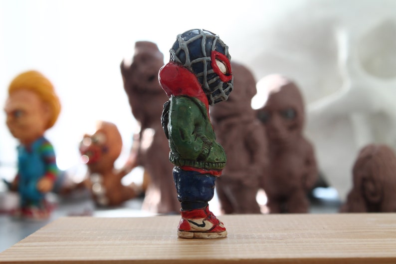 Puede incluir: Una peque&ntilde;a figura de un superh&eacute;roe de color verde, rojo y azul que lleva una m&aacute;scara negra con un dise&ntilde;o de telara&ntilde;a. La figura est&aacute; de pie sobre una superficie de madera.
