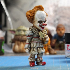 Puede incluir: Una peque&ntilde;a figura detallada de Pennywise el payaso danzante de la pel&iacute;cula "IT". La figura est&aacute; pintada con un estilo realista con una cara blanca, labios rojos y cabello naranja. El payaso lleva un traje blanco y rojo con un cuello con volantes y una corbata de lazo roja. La figura sostiene un peque&ntilde;o objeto blanco en su mano derecha.