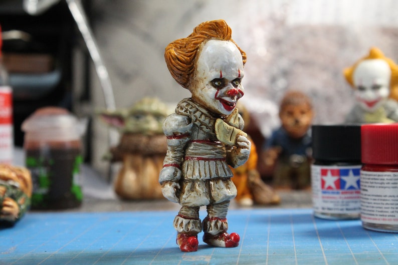 Puede incluir: Una peque&ntilde;a figura detallada de Pennywise el payaso danzante de la pel&iacute;cula "IT". La figura est&aacute; pintada con un estilo realista con una cara blanca, labios rojos y cabello naranja. El payaso lleva un traje blanco y rojo con un cuello con volantes y una corbata de lazo roja. La figura sostiene un peque&ntilde;o objeto blanco en su mano derecha.