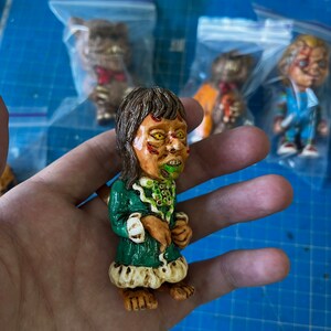 May include: A green and white hand-painted figurine of a character with long brown hair, a green dress, and a green and white face. The figurine is holding a small object in its hand.