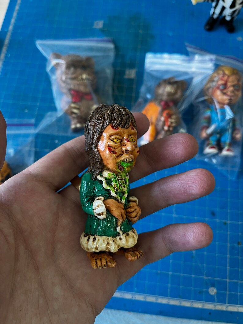 May include: A green and white hand-painted figurine of a character with long brown hair, a green dress, and a green and white face. The figurine is holding a small object in its hand.