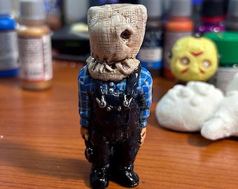 May include: A small, hand-painted figurine of a character wearing a burlap sack over their head, a blue and black plaid shirt, and black overalls. The figurine is standing on a wooden surface.