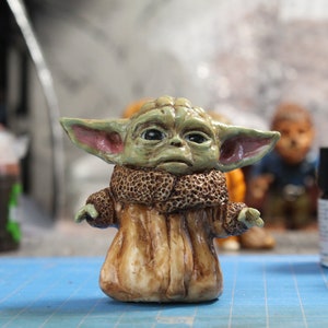 Puede incluir: Una peque&ntilde;a figura verde y marr&oacute;n de un personaje de la franquicia Star Wars. El personaje tiene orejas grandes, ojos grandes y lleva una t&uacute;nica marr&oacute;n. La figura est&aacute; sentada sobre una superficie azul.