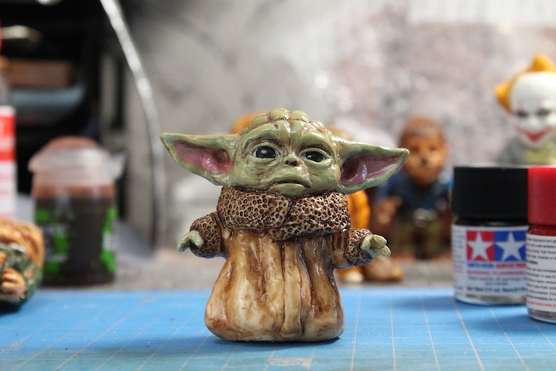 Puede incluir: Una peque&ntilde;a figura verde y marr&oacute;n de un personaje de la franquicia Star Wars. El personaje tiene orejas grandes, ojos grandes y lleva una t&uacute;nica marr&oacute;n. La figura est&aacute; sentada sobre una superficie azul.