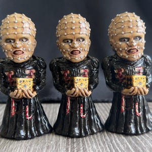 Puede incluir: Tres pequeñas figuras de un personaje con la cabeza cubierta por una rejilla de púas y una túnica negra, cada una sosteniendo una pequeña caja dorada.