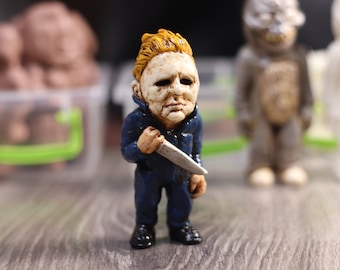 May include: A small, blue-suited figurine of a horror movie character with a white mask and a knife. The figurine has a yellow-blonde hair and a white mask with a dark, painted face.