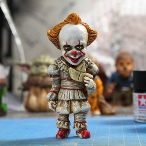 Puede incluir: Una figurita pintada a mano de Pennywise el payaso danzante de la pel&iacute;cula "IT". La figurita lleva un traje blanco y rojo con un barco de papel en la mano. La figurita est&aacute; de pie sobre una superficie azul.
